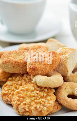 closeup of an assortment of different shortbread biscuits in a white plate, on a set table with some cups of tea or coffee Stock Photo