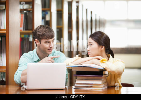 Two college students talking with notebook computer and stacked books at table in library Stock Photo