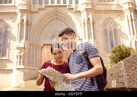 Young tourist couple on vacation with a backpack, looking at a map while dressed casually in jeans and t-shirts with a cathedral Stock Photo