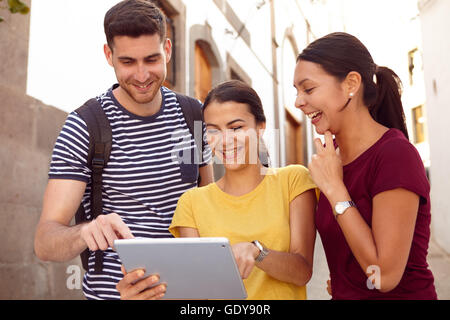 Young tourists on vacation having fun with a backpack looking at a tablet with a friend in a narrow street while dressed in casu Stock Photo