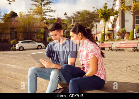 Young couple sitting in a town square on a bench in the sunshine looking at a tablet while dressed casually in jeans and t-shirt Stock Photo