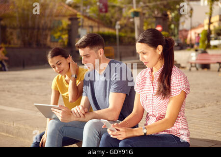 Three young people sitting in a town square in the sun looking at a tablet and cell phone while dressed casually Stock Photo