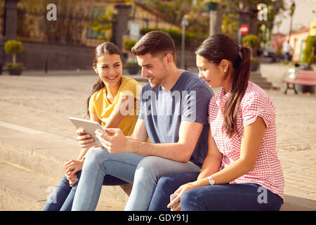 Three laughing young people sitting in a town square on a step looking at a tablet while dressed casually in jeans and t-shirts Stock Photo