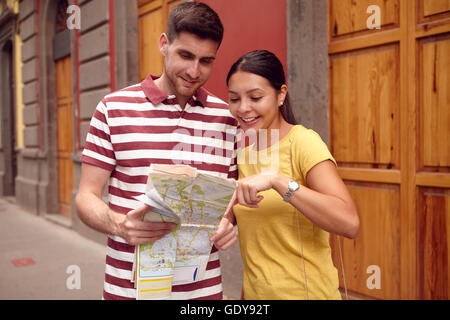 Young couple studying a map and pointing at it while smiling happily and dressed casually in t-shirts with old buildings behind  Stock Photo