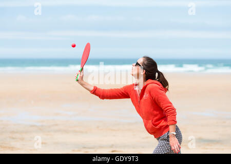 A young woman playing a bat and ball game on a beach wearing casula clothes in summer UK Stock Photo