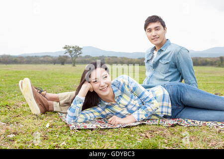 Young smiling woman lying side down and young smiling man sitting on grassland staring at front Stock Photo