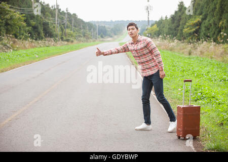 Young man standing and trying to hitch a ride with his carrier on a road Stock Photo