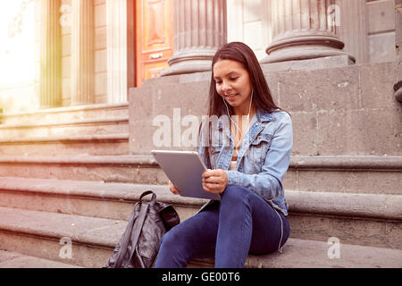 Pretty young girl looking at her tablet while sitting on the steps of an old building smiling wearing her hair loose and casual  Stock Photo
