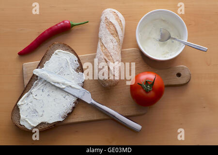 Cheese spreading on slice of brown bread with baguette and tomato on chopping board Stock Photo