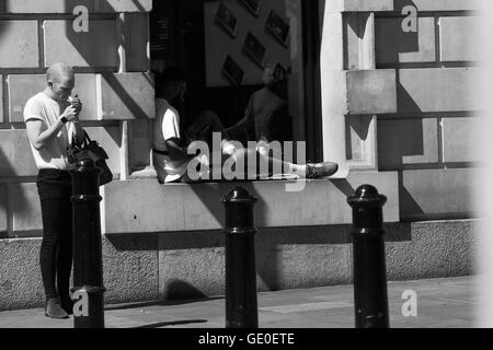 In Covent Garden a man sits in shadow on a window ledge while another stands close by lighting a cigarette on a hot summer day. Stock Photo