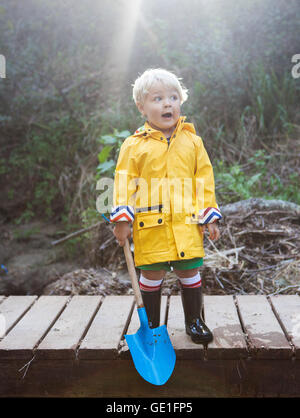 Toddler boy in a raincoat standing on wooden jetty holding a shovel Stock Photo