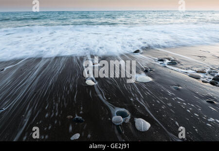 Pebbles on  the beach on a black sand and flowing sea water creating nice textures. Long exposure image. Stock Photo