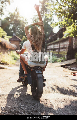 Rear view shot of young couple riding motorcycle on rural road. Young female sitting on back of her boyfriend riding bike. Stock Photo