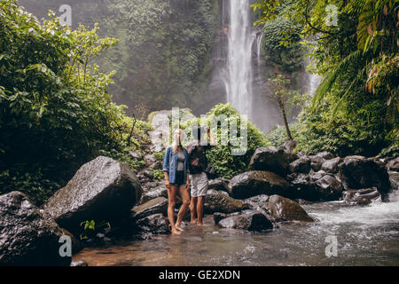 Young couple standing by the stream, with man taking photographs of waterfall. Couple of tourist enjoying in nature. Stock Photo