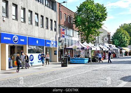 Shoppers in Brentwood Essex shopping High Street Boots pharmacy store and canopies above street market stalls beside cobblestone road England UK Stock Photo