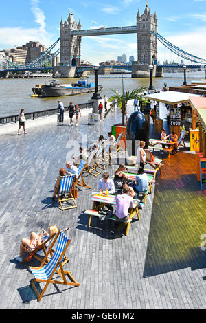 Tourists & office workers lunch time at fast food stalls set up for summertime alfresco snacks deckchairs beside River Thames Tower Bridge UK beyond Stock Photo
