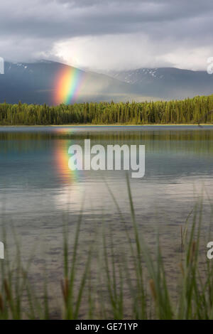 Rainbow emerges from gap in clouds and is reflected in Boya Lake, BC Stock Photo