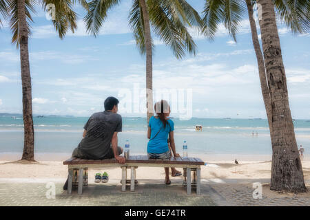 Pattaya beach Thailand. Couple sat on a bench amongst palm trees and looking out over the sea. Pattaya Thailand S. E. Asia. Stock Photo