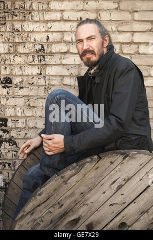 Outdoor portrait of young bearded Asian man sitting on old wooden cable reel Stock Photo