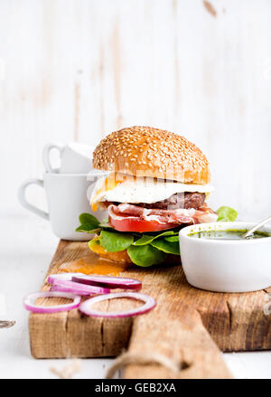 Breakfast set. Homemade beef burger with fried egg, vegetables, onion rings and coffee cups on wooden board Stock Photo