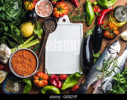Ingredients for cooking healthy dinner. Raw uncooked seabass fish with vegetables, grains, herbs and spices over rustic wooden background, white ceramic board in center Stock Photo