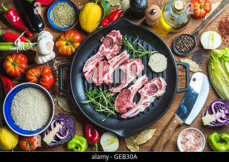 Ingredients for cooking healthy meat dinner. Raw uncooked lamb chops in iron grill pan with vegetables, rice, herbs and spices over rustic wooden background Stock Photo