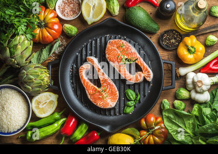 Dinner cooking ingredints. Raw uncooked salmon fish with vegetables, rice, herbs, spices and oil in iron grilling pan over rustic wooden background Stock Photo