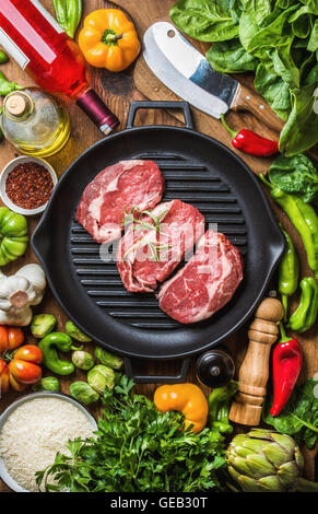 Ingredients for cooking healthy meat dinner. Raw uncooked beef steaks with vegetables, rice, herbs, spices and wine bottle over rustic wooden background, Stock Photo