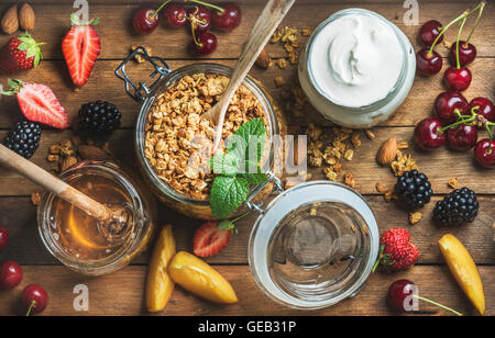 Healthy breakfast ingredients. Oat granola in open glass jar, fruit, yogurt, berries and mint over wooden background Stock Photo