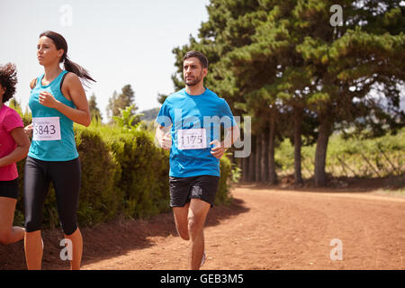 Three marathon runners in a race on a gravel path with trees behind them wearing casual running clothes in the late morning sun Stock Photo