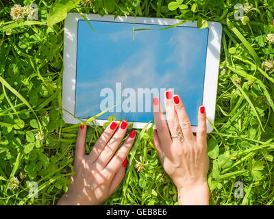 Beautiful female hands with red nails using white tablet PC in green grass outdoors. Sky and grass reflection in the screen Stock Photo
