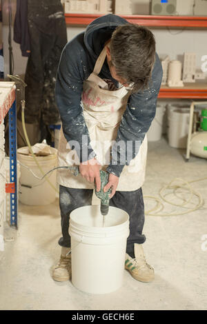 A man in plaster-splattered clothing stirring contents of a white plastic bucket using a mixer attached to an electric drill Stock Photo