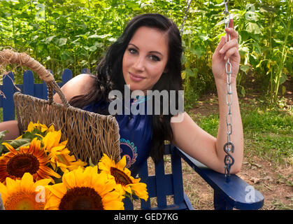 Pretty, young, brunette woman sitting on swing holding bright yellow sunflowers Stock Photo