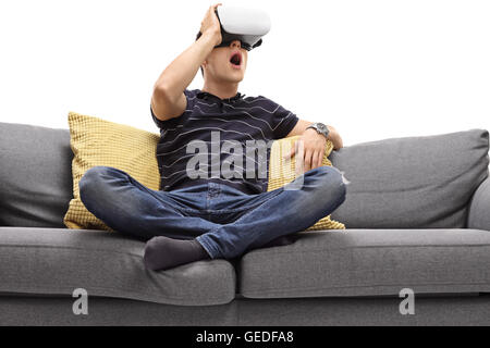 Excited young man looking in VR goggles seated on a gray sofa isolated on white background Stock Photo