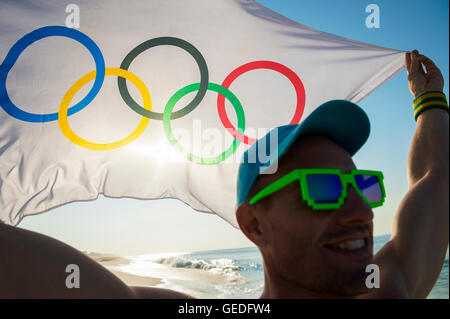 RIO DE JANEIRO - MARCH 10, 2016: Smiling Brazilian athlete holds Olympic flag at sunrise view over the beach shore. Stock Photo