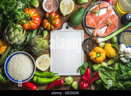 Raw uncooked salmon fish in small metal plate with vegetables, rice, herbs, spices, olive oil on rustic wooden background, white ceramic board in center, copy space, top view Stock Photo