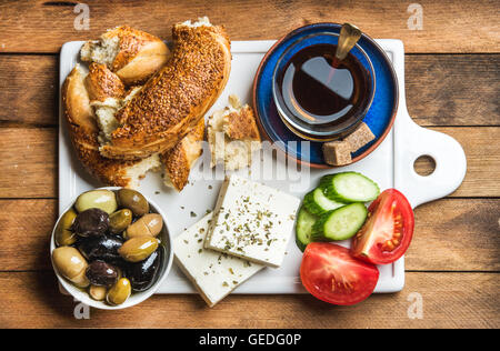 Turkish traditional breakfast with feta cheese, vegetables, olives, simit bagel and black tea on white ceramic board over wooden background. Top view Stock Photo