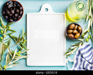 Two bowls with pickled green and black olives, olive tree sprigs, oil in glass bottle, white ceramic board in center over blue background, copy space Stock Photo