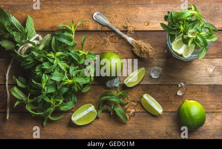 Ingredients for making mojito summer cocktail. Fresh mint bunch and leaves, limes, spoon of brown sugar, ice over rustic wooden background, top view Stock Photo