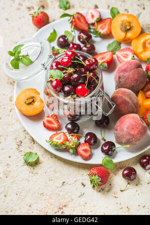 Healthy summer fruit variety. Sweet cherries in glass jar, strawberries, peaches, apricots and mint leaves on white ceramic serving plate over light concrete background. Top view, selective focus Stock Photo