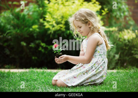 Little blonde girl holding young flower plant in hands on green background Stock Photo