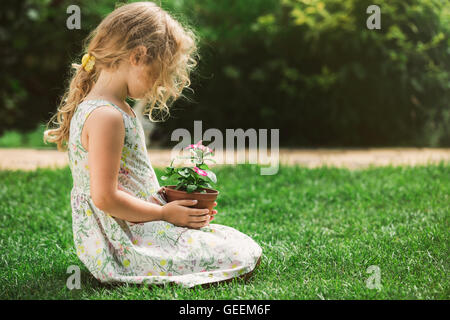 Little blonde girl holding young flower plant in hands on green background Stock Photo