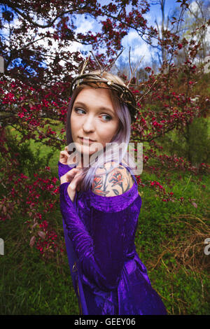 Young woman dressed in a purple velvet gown and twig crown with flowering crab apples in the background Stock Photo