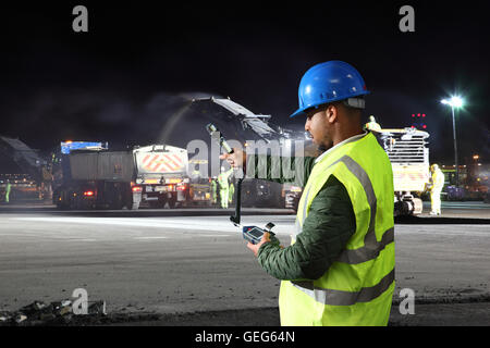 An environmental engineer monitors dust levels next to road resurfacing machines working overnight at a London airport. Stock Photo