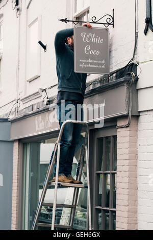 A man on a ladder fixing a painted name sign onto a bracket on a shopfront. Stock Photo