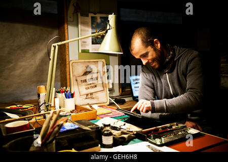 A man in his workshop using a digital tablet with a touch screen surrounded by equipment for signwriting and screen-printing. Stock Photo