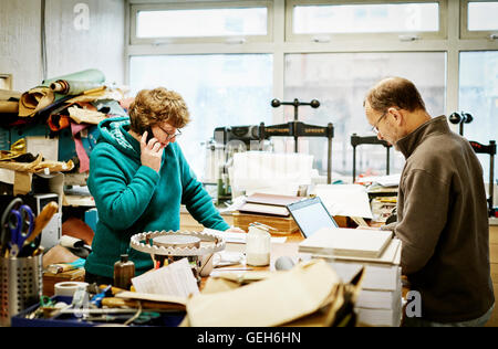 A woman taking a phone call and a man working on a laptop computer in a bookbinding workshop. Stock Photo