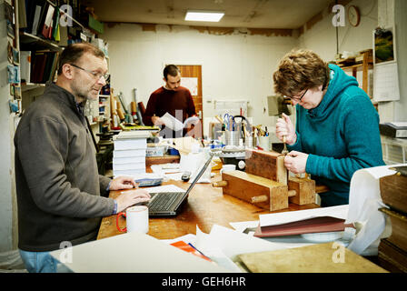 A woman working on the spine of a bound book with a hand tool. A man using a laptop computer. Stock Photo