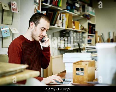 A man using a smart phone and digital tablet in a small bookbinding workshop. Stock Photo