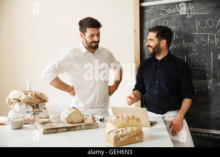 Two bakers standing at a table, using a laptop computer, freshly baked bread, a blackboard on the wall. Stock Photo